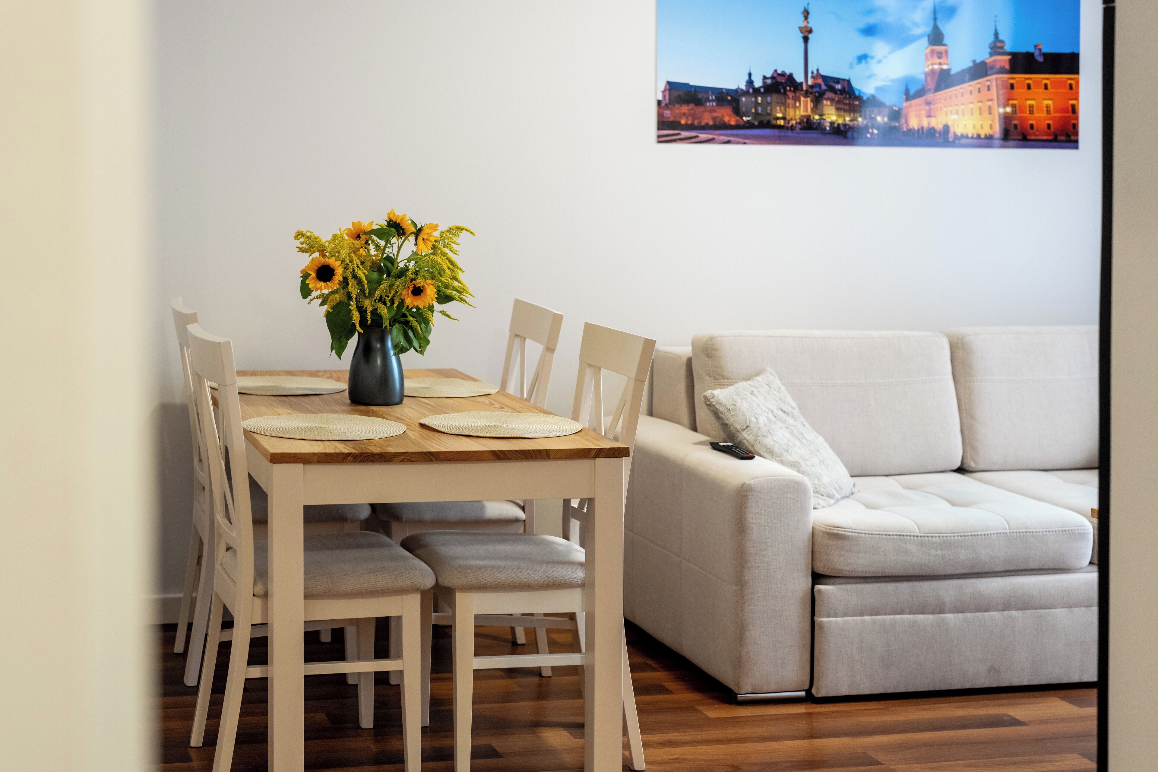 Un coin repas avec une table en bois, des chaises blanches et un vase rempli de tournesols. Un canapé beige est placé contre le mur, et un tableau représentant un paysage urbain, encadré, est accroché au-dessus du canapé.