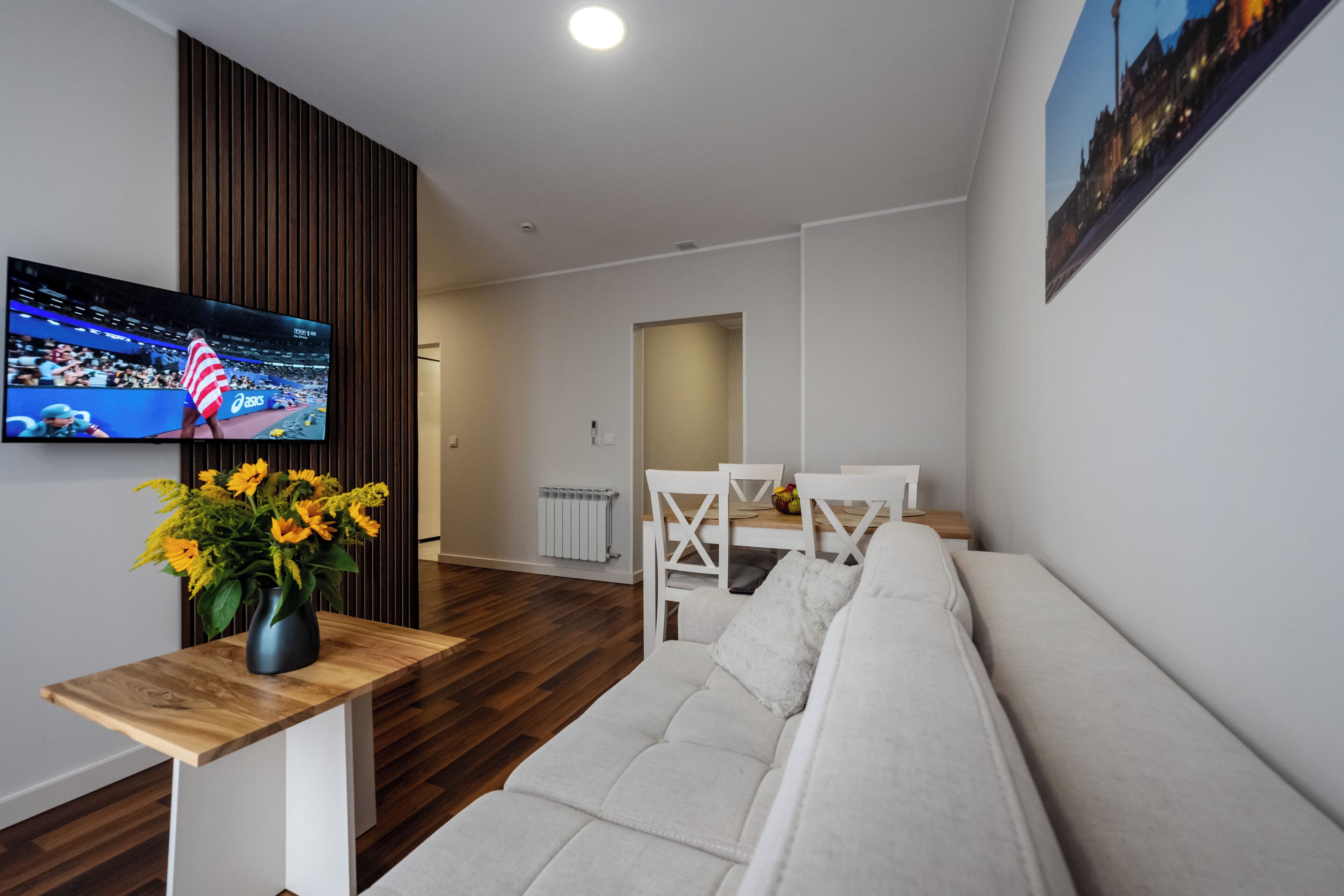 A modern living room with a flat-screen TV, a wooden coffee table, a white sofa, and a vase of sunflowers.