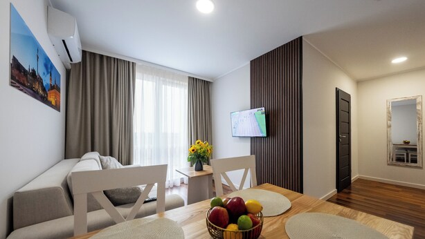 A modern living room with a wooden dining table, white chairs, a TV, and a fruit bowl. - Wesoła Apartamenty (Wólka Kosowska)