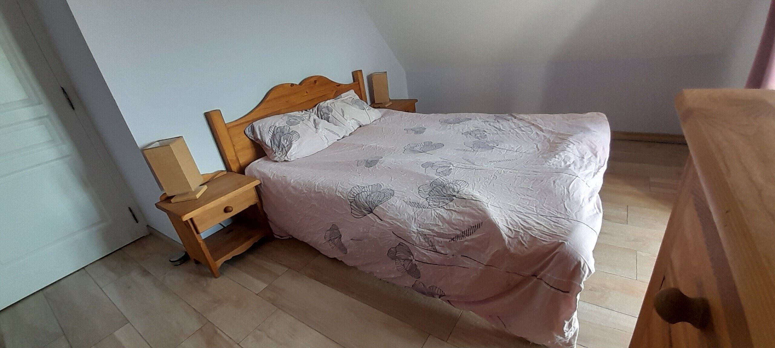 Uma cama de casal com cabeceira e peseira de madeira, um criado-mudo e uma mesinha de cabeceira.