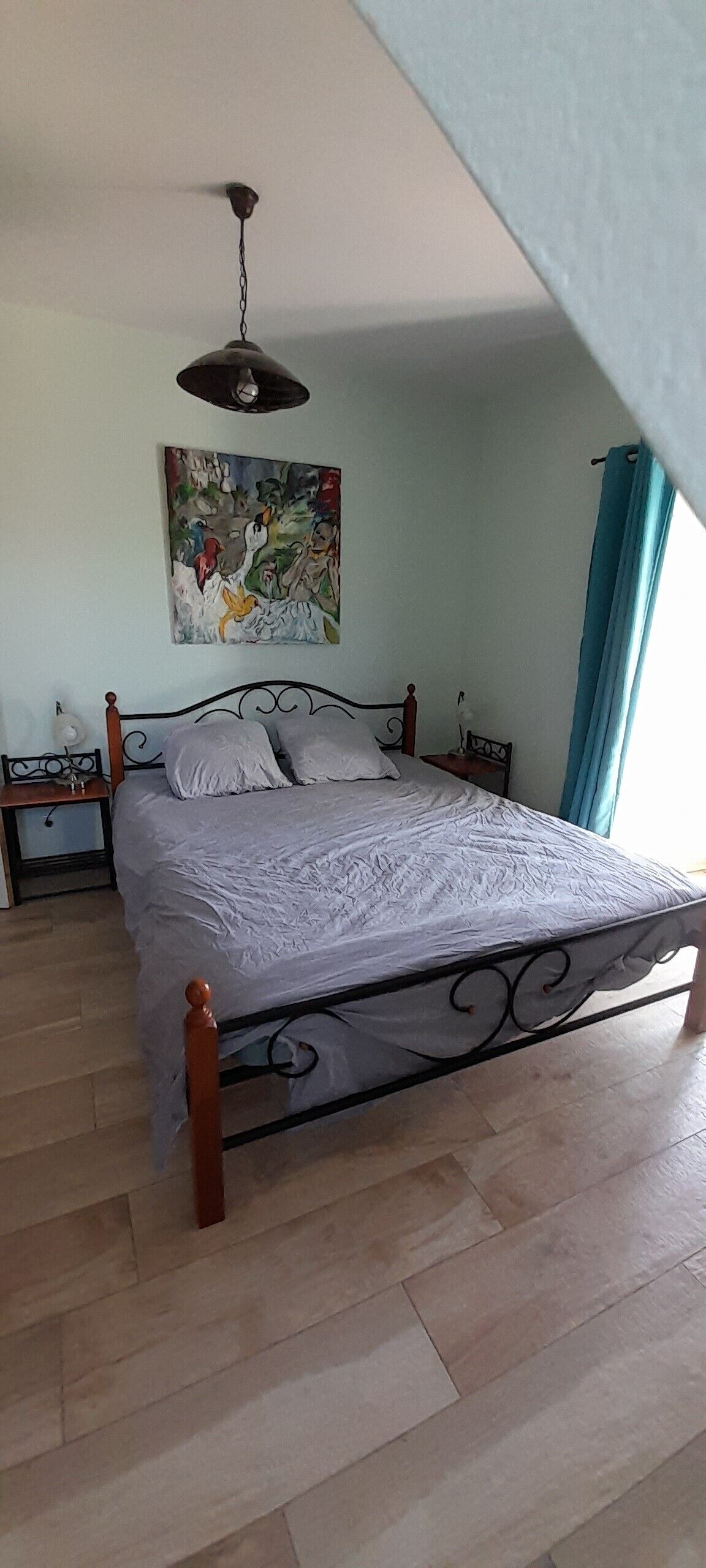 A bedroom with a bed, a nightstand, a lamp, a ceiling fan, a painting, and a window with curtains.