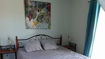 A bedroom with a bed, a nightstand, a lamp, a ceiling fan, a painting, and a window with curtains.
