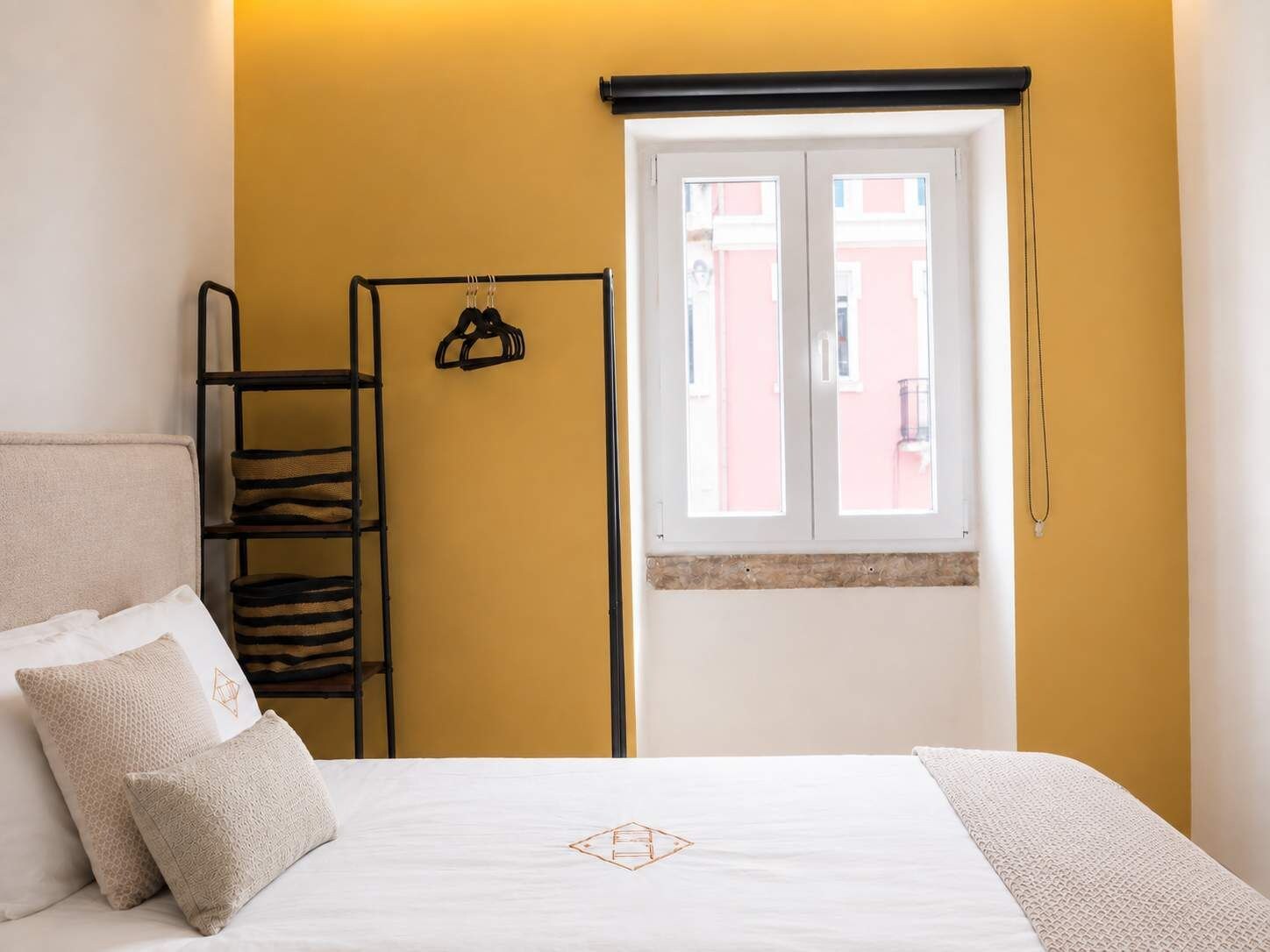 Una habitación de hotel con una cama, un ventanal con vistas a edificios, un perchero metálico negro con objetos y una pared blanca.