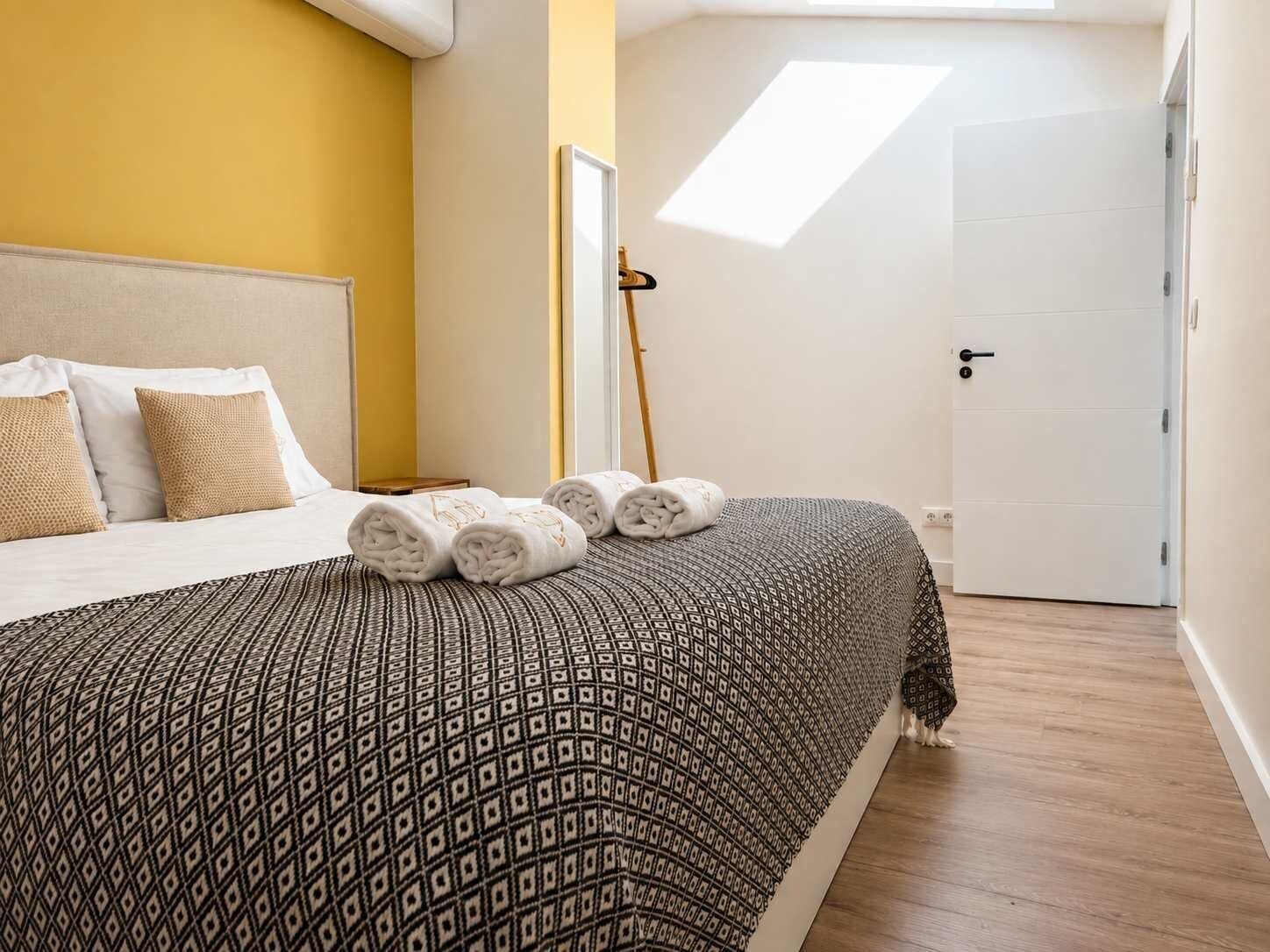 Un dormitorio con cama, almohadas, toallas enrolladas, colcha estampada, suelo de madera, puerta blanca y tragaluz.