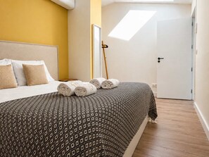 A bedroom with a bed, pillows, rolled towels, a patterned bedspread, a wooden floor, a white door, and a skylight. - Spacious Gulbenkian – Families, A/C & Remote Work (Lisboa)