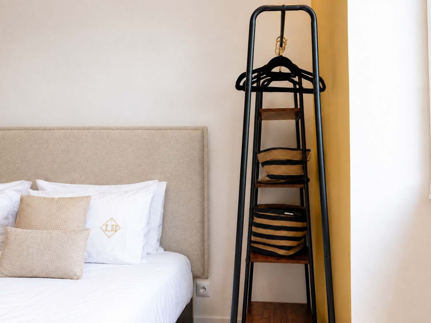 A bed with white bedding and a headboard, a black metal shelf unit with baskets, and a wall socket.