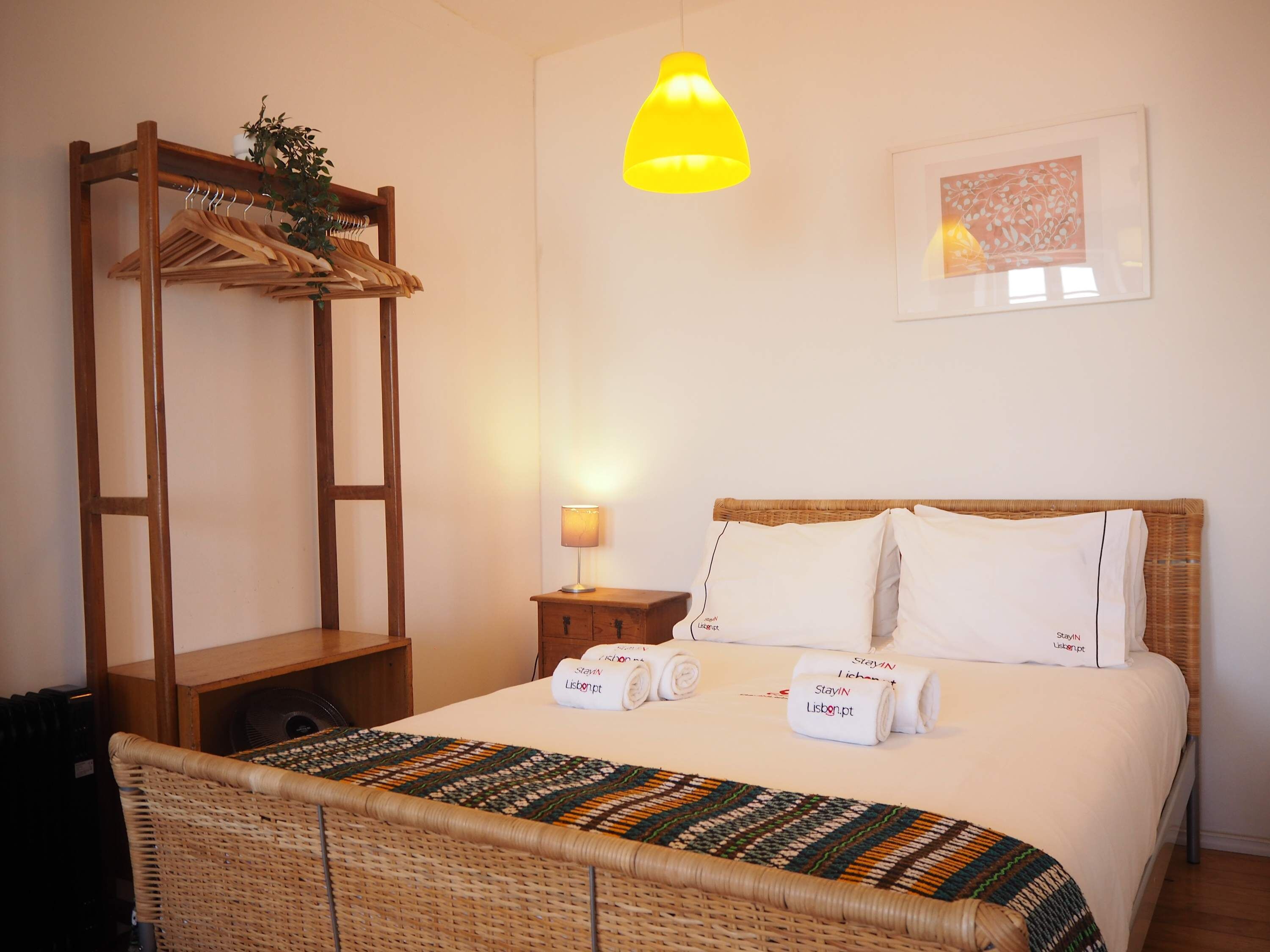 Une pièce comprenant un lit, une armoire en bois, une plante suspendue, un luminaire suspendu jaune et un tableau encadré au mur.