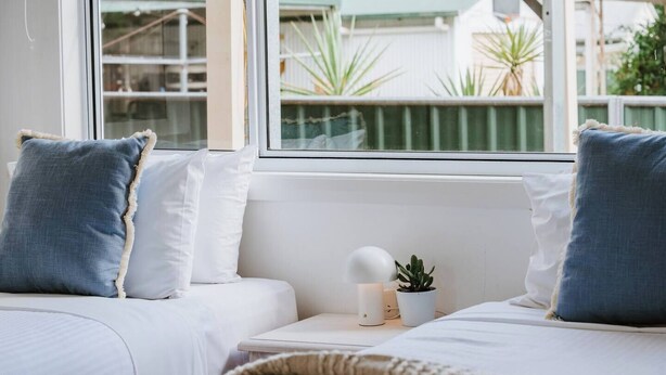 Un dormitorio con dos camas, ropa de cama blanca, un ventanal con vista a zonas verdes y una mesita de noche con una lámpara y una planta.