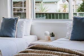 Une chambre avec deux lits, du linge de lit blanc, une fenêtre donnant sur un paysage verdoyant, et une table de chevet avec une lampe et une plante.