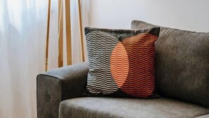 Un sofá gris con cojines estampados, una lámpara de pie y una cortina blanca en una habitación con piso de madera.