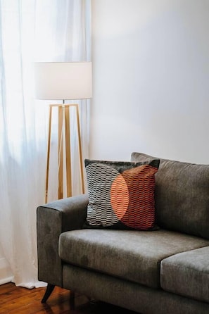 A grey sofa with patterned cushions, a floor lamp, and a white curtain in a room with wooden flooring.