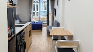 Uma cozinha compacta com máquina de lavar, área de jantar com mesa e cadeiras, cama com colcha azul e vista para um edifício pela janela.