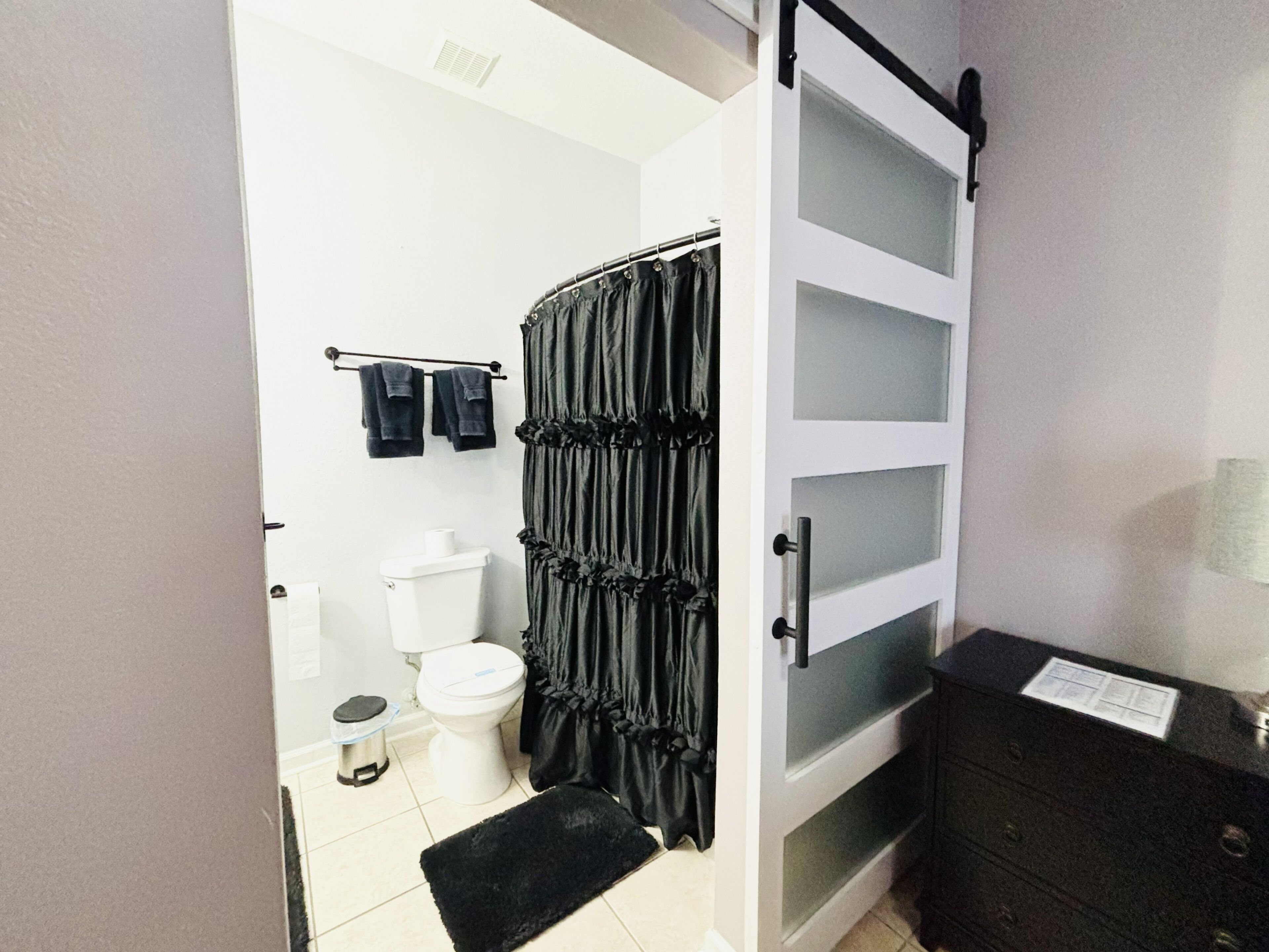 A bathroom with a toilet, a black shower curtain, a towel rack with two towels, a black bench, and a cabinet with a paper on top.