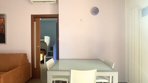 A room with a glass-top table, white chairs, a brown sofa, a clock, and an air conditioning unit. - PP08 - Porto Potenza, two-room apartment with garden, 100 meters from the sea (Potenza Picena)