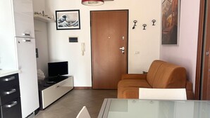 A compact living space with a kitchenette, a sofa, a glass table, and artwork on the walls. - PP08 - Porto Potenza, two-room apartment with garden, 100 meters from the sea (Potenza Picena)