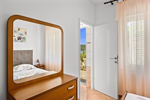 Une chambre avec une commode en bois, un grand miroir, un lit recouvert d’une couvre-lit à motifs floraux, une table de chevet avec une lampe et une porte donnant sur un balcon.