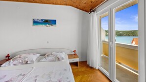 A bedroom with a wooden ceiling, a bed with floral bedding, a nightstand with a lamp, a painting of a beach scene, and a balcony with a view of the sea. - Two Bedroom Apt near the Beach - Villa Jagoda (Kampor)