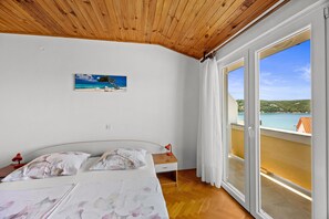 Ein Schlafzimmer mit Holzboden, einem Bett mit floralem Bettzeug, einem Nachttisch mit Lampe, einem Gemälde mit Strandmotiv und einem Balkon mit Meerblick.