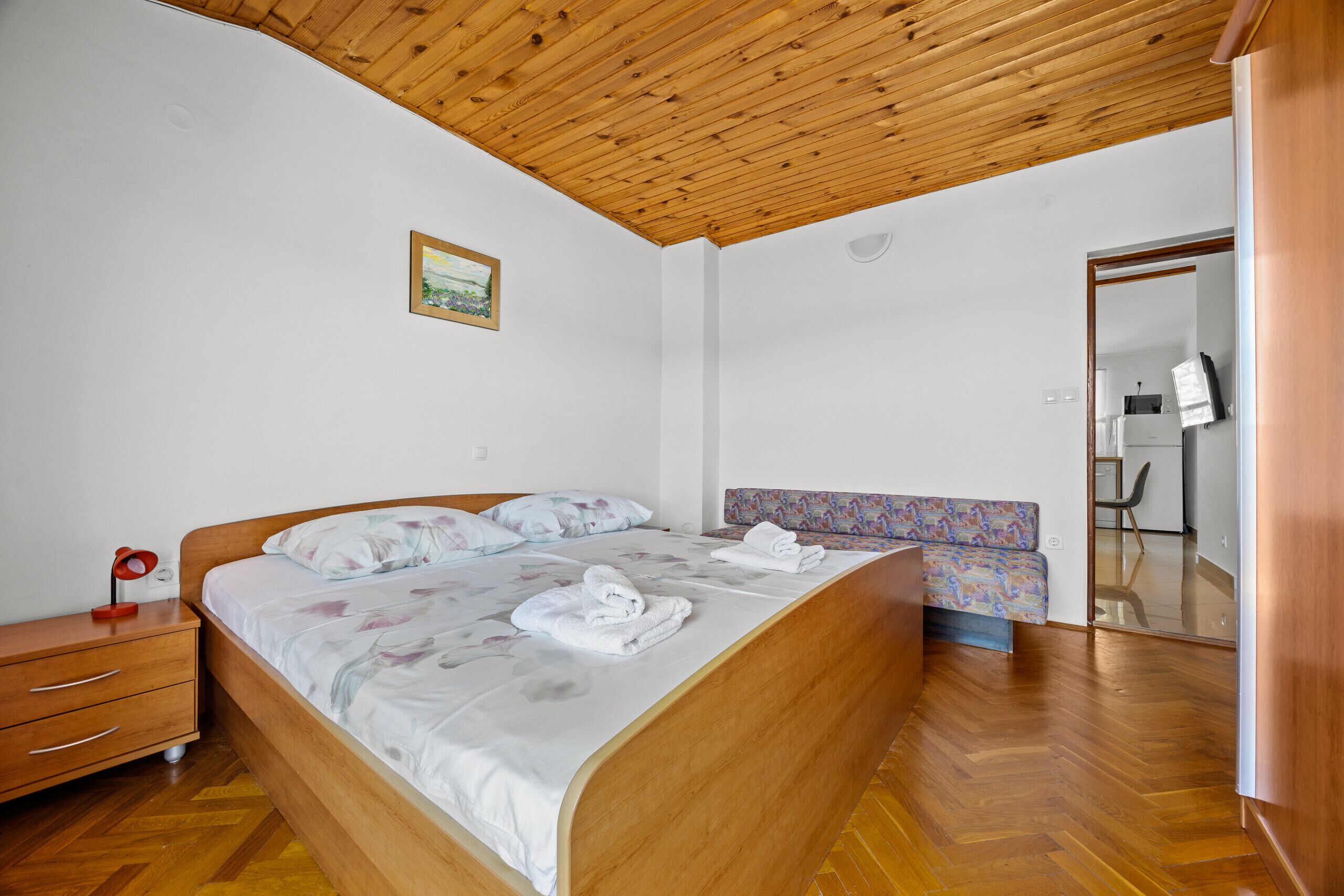 A bedroom with a wooden ceiling, a bed with floral bedding, a bedside table, and a mirror.