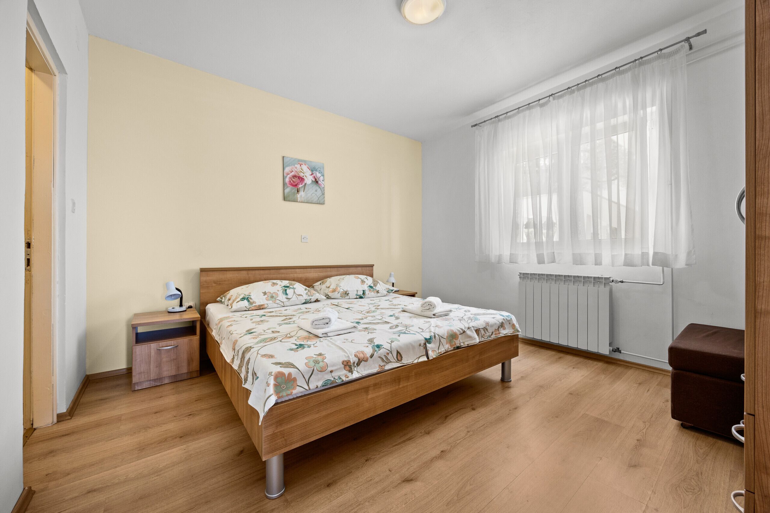 Un dormitorio con cama de madera, mesita de noche, radiador y ventana con cortinas translúcidas.