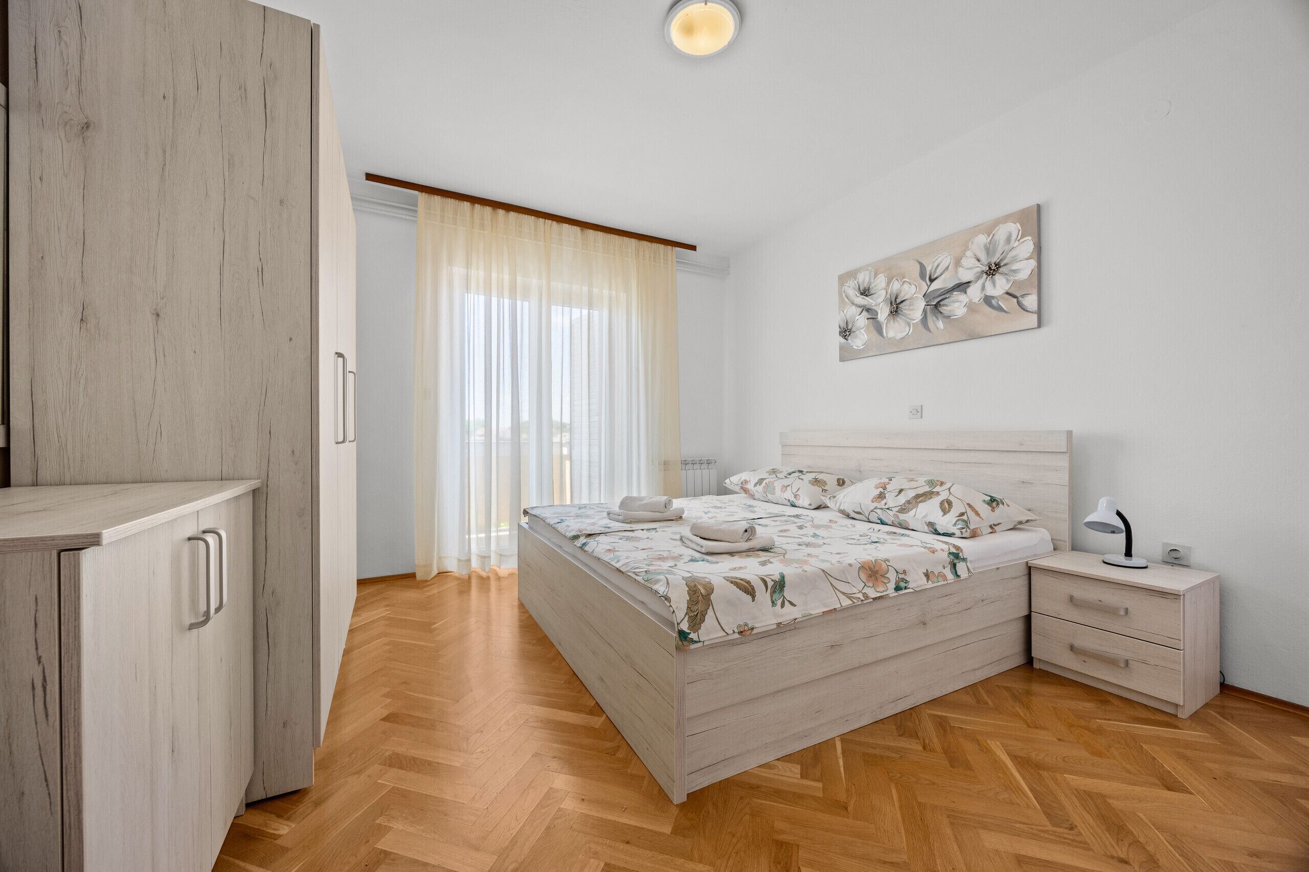 Ein Schlafzimmer mit einem Holzschrank, einem Bett mit floralem Bettzeug, einem Nachttisch und einem an der Wand befestigten Kunstwerk.