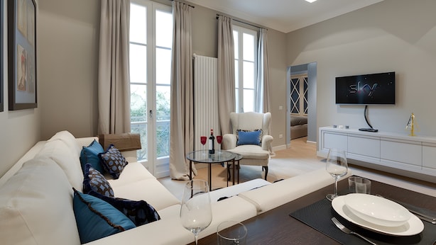 Ein modernes Wohnzimmer mit einer Couch, einem Glastisch, einem Essbereich mit weißem Tisch und Tellern, einem Fernseher und großen Fenstern mit Vorhängen.
