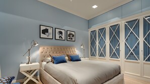 Ein Schlafzimmer mit einer gesteppten Kopfteil, einem großen Bett, zwei Nachttischlampen, einem Nachttisch, einem blauen Sessel und einem Kleiderschrank mit geometrischem Fensterdesign.