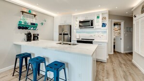 A modern kitchen with white cabinets, a marble island, blue bar stools, and a microwave. - NEW | Beachfront | Heated Pools | Hot Tub| Balcony|Ocean Views | Patio | Grills | Cabanas | Pets OK (Galveston)