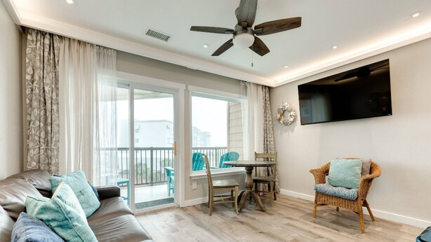 A living room with a sofa, a dining table, chairs, a ceiling fan, and a TV mounted on the wall. - NEW | Beachfront | Heated Pools | Hot Tub| Balcony|Ocean Views | Patio | Grills | Cabanas | Pets OK (Galveston)