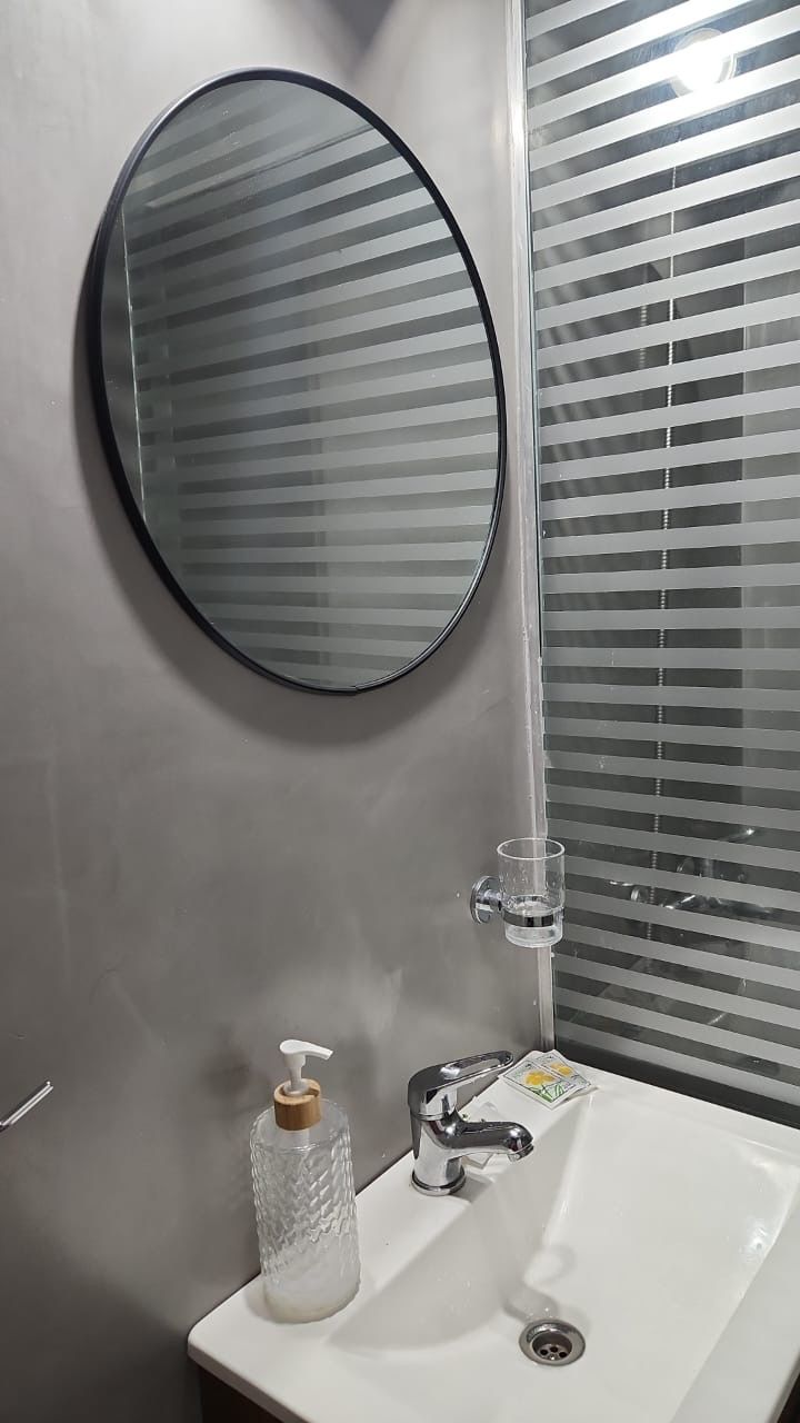 Un baño con un espejo redondo, un lavabo blanco, un dispensador de jabón y un vaso de agua.