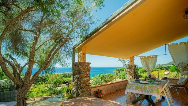 Un'area pranzo all'aperto riparata, con un tavolo apparecchiato per due, affacciata su una vista panoramica del mare e del verde lussureggiante.