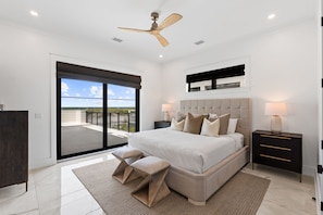 A bedroom with a large bed, a ceiling fan, a nightstand with a lamp, a bench, and a view through a sliding door to an outdoor balcony. - Rumor Has It: Beachfront, Private Pool, Pet Friendly, Golf Cart Community (Mexico Beach)