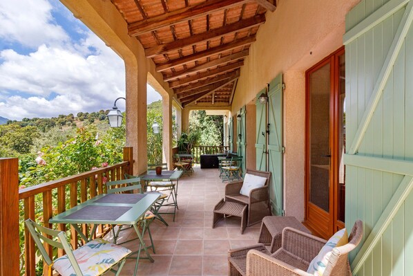 A covered outdoor patio with tables and chairs, a wooden railing, and a view of greenery.