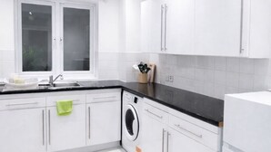 A modern kitchen with white cabinetry, a black countertop, a built-in washing machine, and a window with frosted glass. - Stylist 7sleeps contractor groups friendly1bedroom flat Canada Water free WiFi (London)