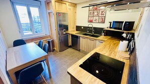 Een kleine keuken met een koelkast, fornuis, magnetron en spoelbak. Er is een eettafel met stoelen en een raam dat natuurlijk licht binnenlaat.