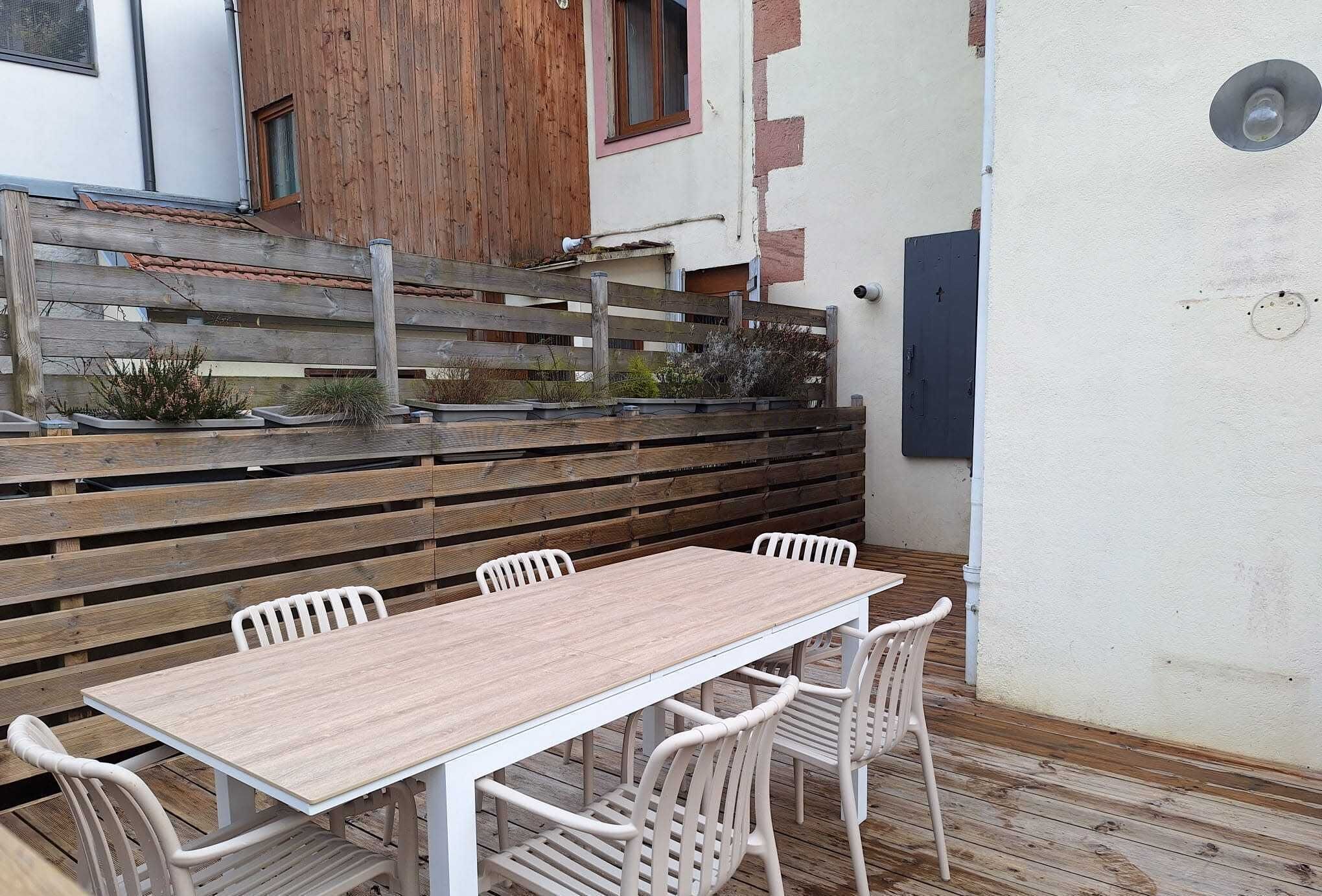 Una terraza de madera con una mesa y sillas blancas, una valla de madera, y un edificio con una ventana y una puerta.