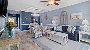 A living room with a ceiling fan, a blue sofa, patterned chairs, a coffee table, and a TV. - Cypress Bend @ Barefoot - Updated w/ Pools, Golf, Shuttle (North Myrtle Beach)