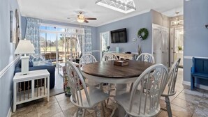 A dining area with a round table, white chairs, a ceiling fan, and a TV mounted on the wall. - Cypress Bend @ Barefoot - Updated w/ Pools, Golf, Shuttle (North Myrtle Beach)