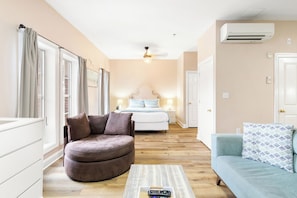Een hotelkamer met een gebogen bruine bank, een lichtblauwe bank, een houten salontafel met een afstandsbediening, een bed met wit beddengoed en een plafondventilator.