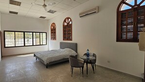 A hotel room with a bed, two chairs, a small table, a ceiling fan, and large windows with wooden shutters.