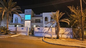 A white building with arched windows and a balcony, surrounded by palm trees and a gated entrance.