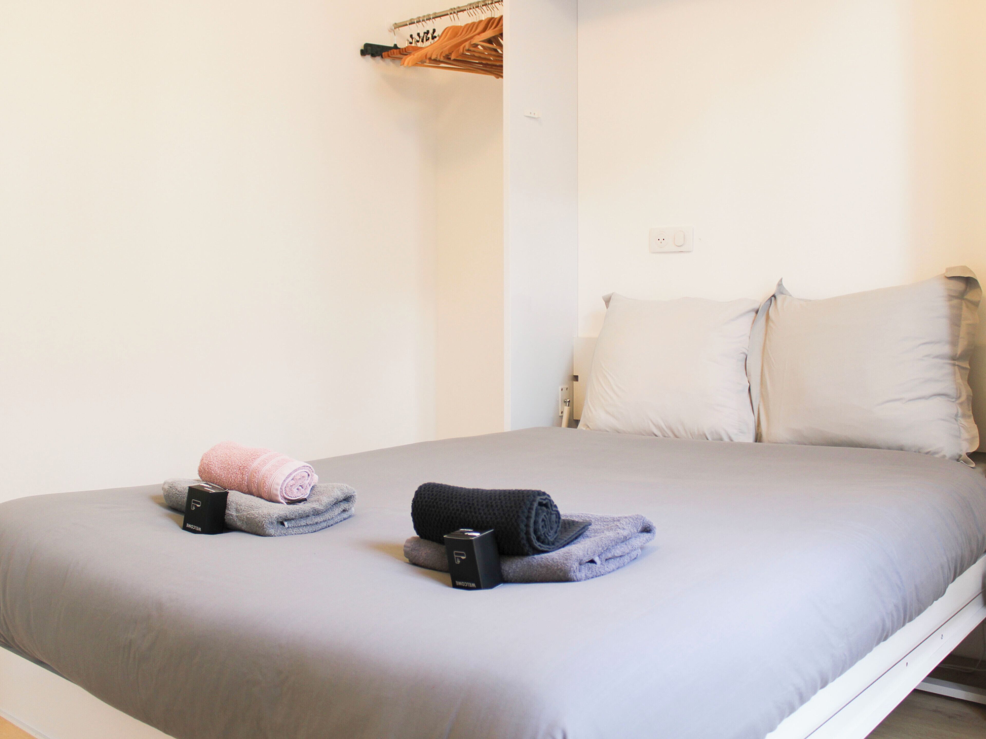 Un lit bien fait, avec un drap-housse gris, deux serviettes enroulées et un objet noir posé sur le lit.