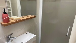 Et badeværelse med en hvid vask, et spejl, en hylde med toiletartikler og en brusekabine i glas.
