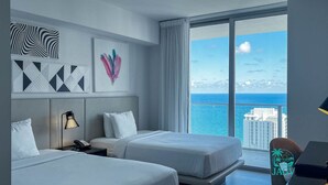A hotel room with two beds, a desk, and a large window with a city and ocean view.