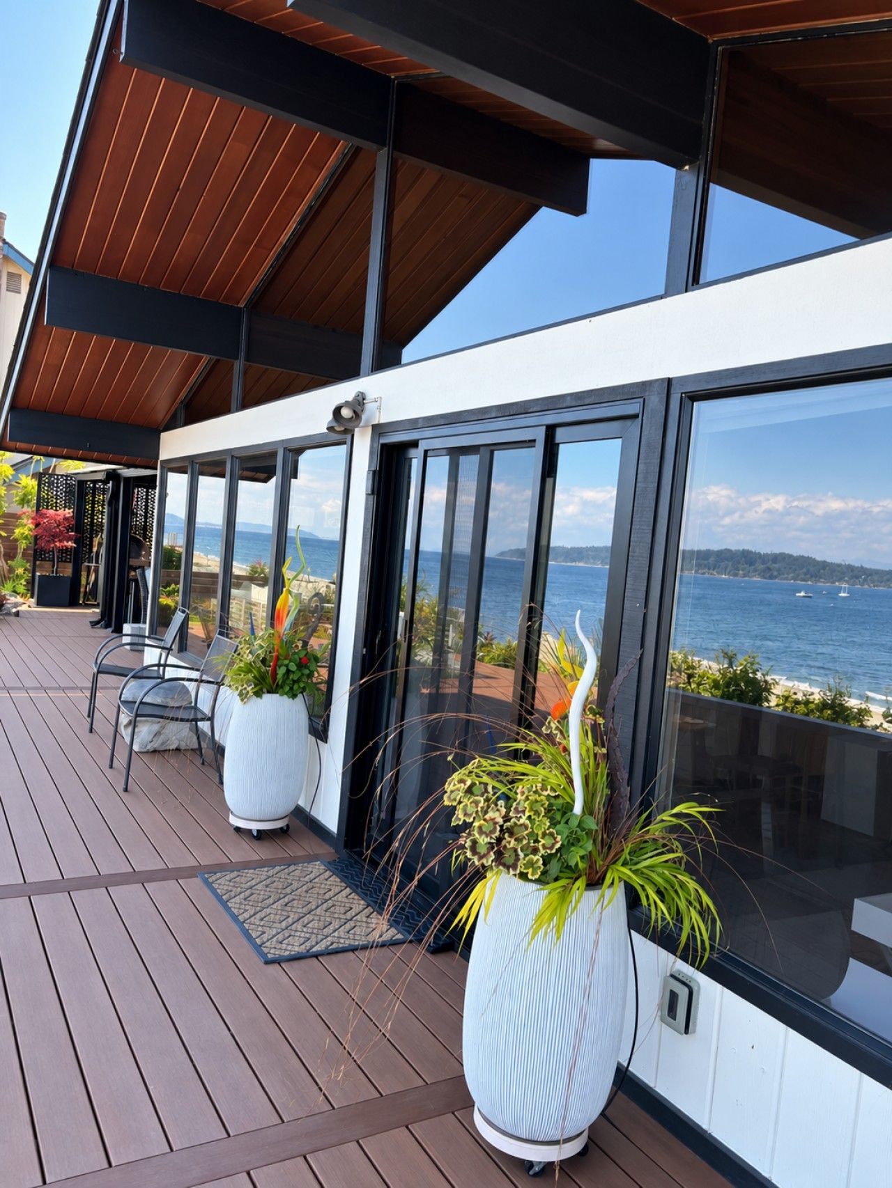A modern outdoor deck with a view of the sea, featuring a seating area with chairs and large potted plants.