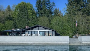 Ein modernes Gebäude direkt am Wasser mit großer Glasfassade, Außenbereich zum Sitzen und einer Fahnenstange.