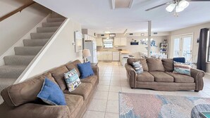 Een ruime woonkamer met een trap, twee bruine banken met decoratieve kussens, een plafondventilator en een keuken op de achtergrond.