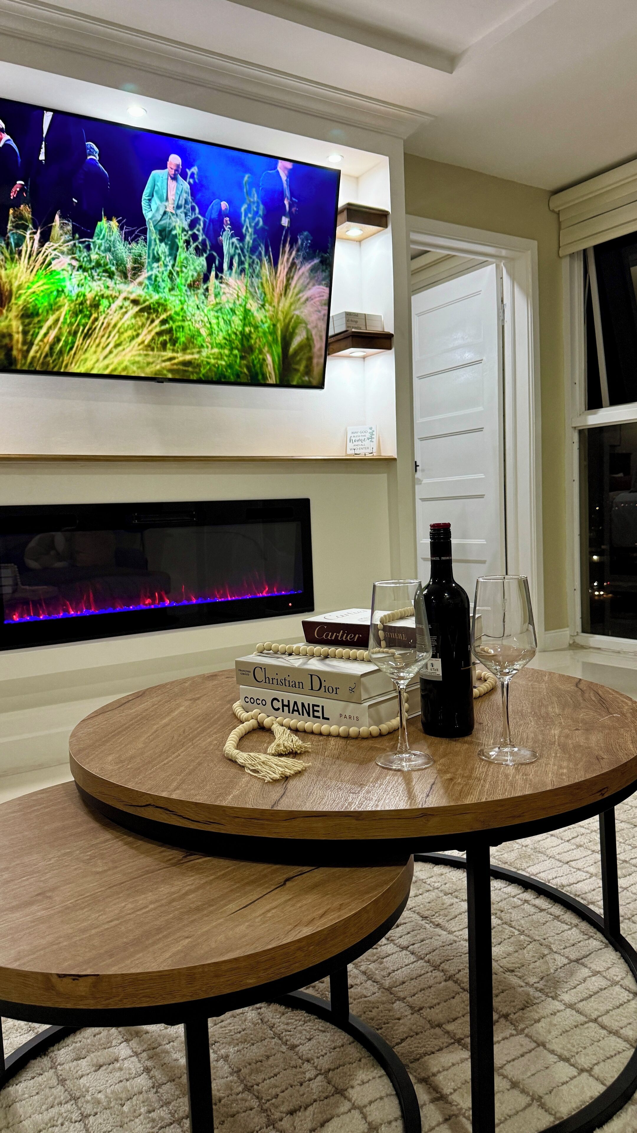 A living room with a flat-screen TV mounted on the wall, a fireplace below, and a round wooden table with a bottle of wine and glasses.