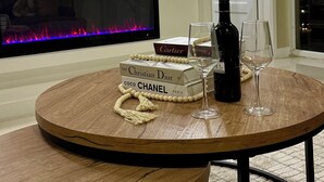 Ein Wohnzimmer mit flachbildfernseher an der Wand, Kamin darunter und einem runden Holztisch mit Weinflasche und Gläsern.
