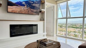 Ein modernes Wohnzimmer mit einem an der Wand montierten Flachbildfernseher, einem Kamin darunter und einem großen Fenster mit Blick auf die Stadt.